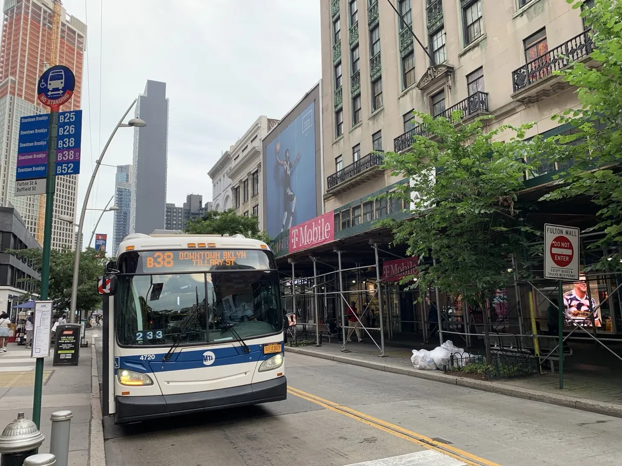 B38 bus at a stop on the car-free Fulton Mall in Downtown Brooklyn. The bus has a white livery with a blue stripe across the front with the MTA logo in white. A sign shows this bus stop is Fulton St & Duffield St and the B25, B26, B38 Limited, and B52 also stop here.